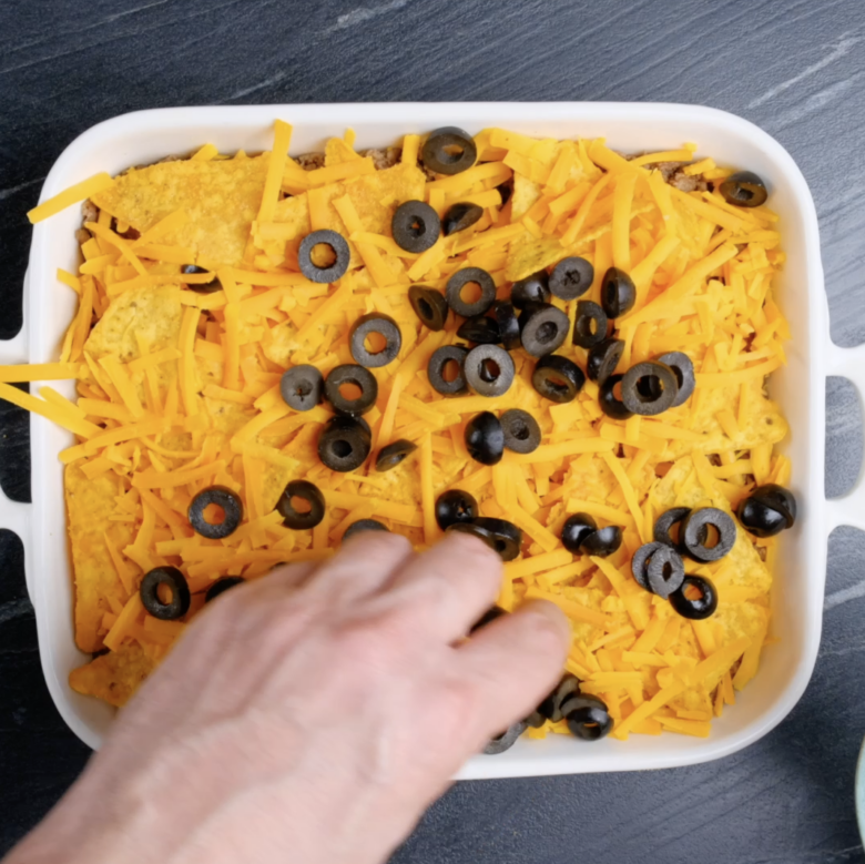 Taco casserole being topped with cheese and black olives before baking.