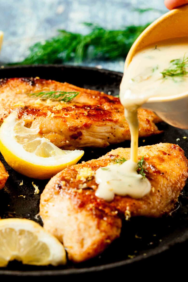 Creamy lemon sauce being poured over skillet chicken with fresh dill in the background.