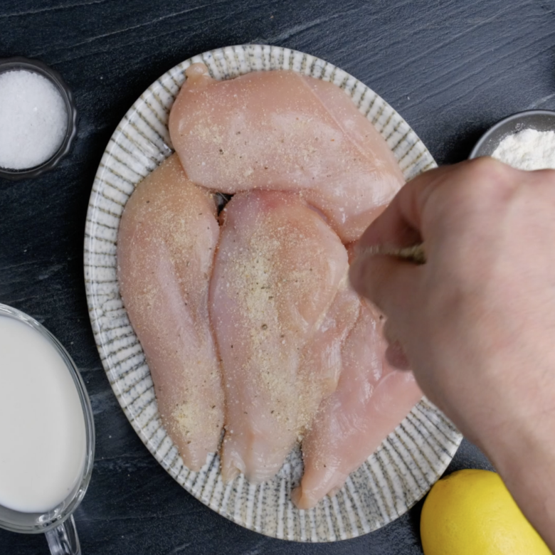 Chicken breast on a white plate being seasoned.