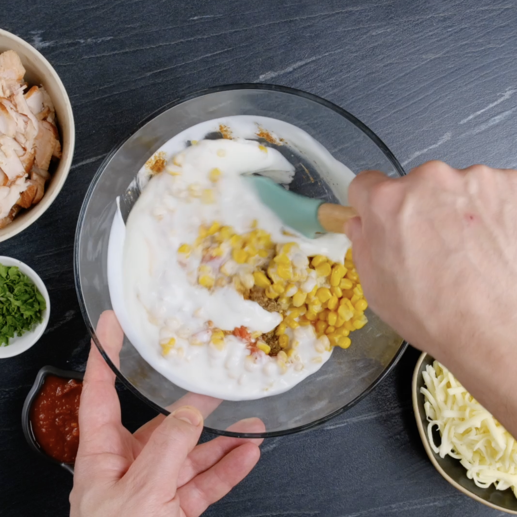 Yogurt being mixed with corn, tomatoes with chiles, and spices to make enchilada mixture.