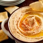 A bowl of hummus garnished with paprika and sesame seeds with crackers next to it.