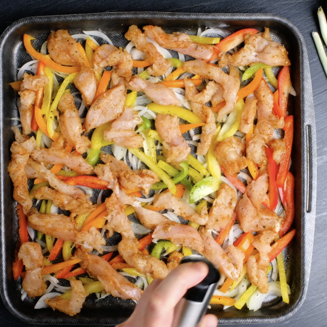 Sheet pan with sliced onions, bell peppers, and seasoned chicken strips to make sheet pan chicken fajitas.