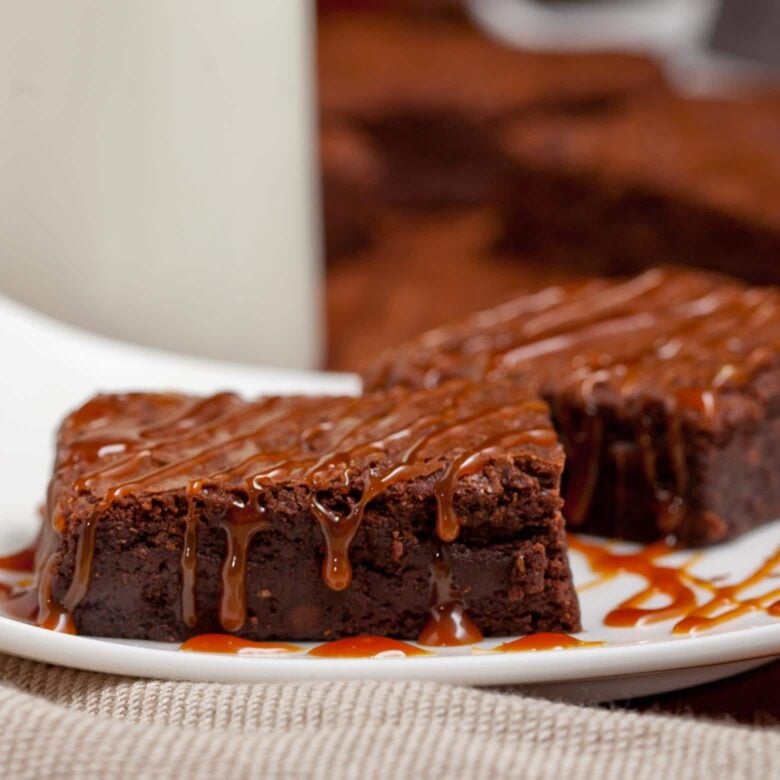 Two fudgy dark chocolate brownies drizzled with salted caramel sauce on a white plate.