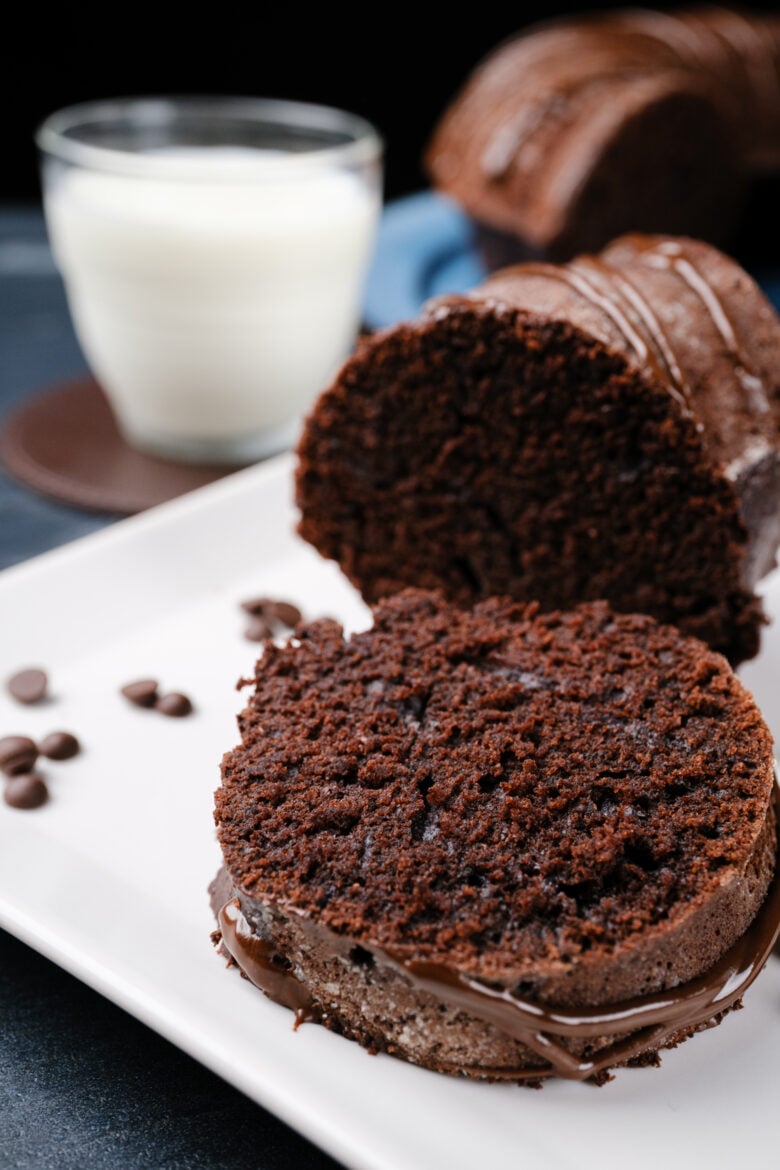 A slice of chocolate bundt cake on a plate next to a glass of milk.