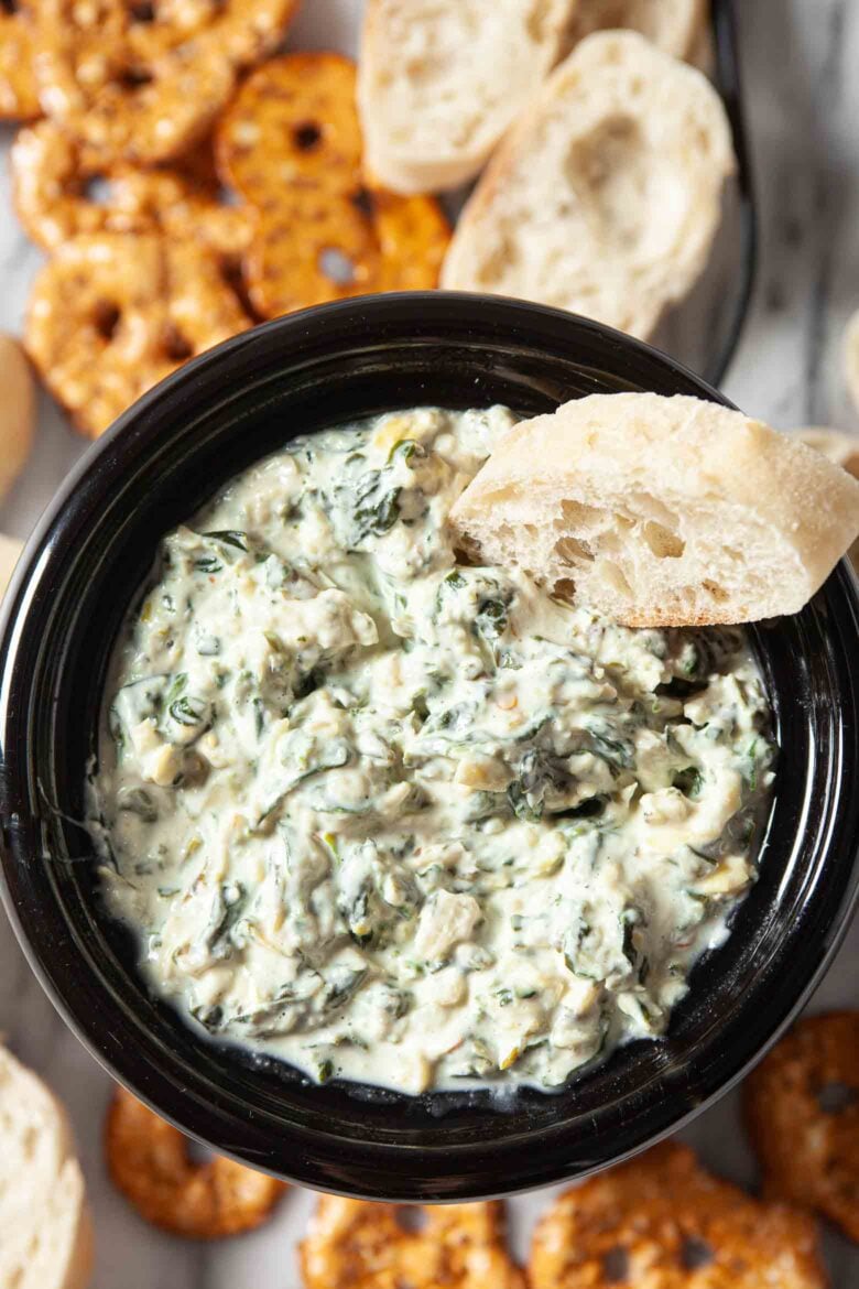 A bowl of crockpot spinach and artichoke dip with pretzels and pretzel sticks.