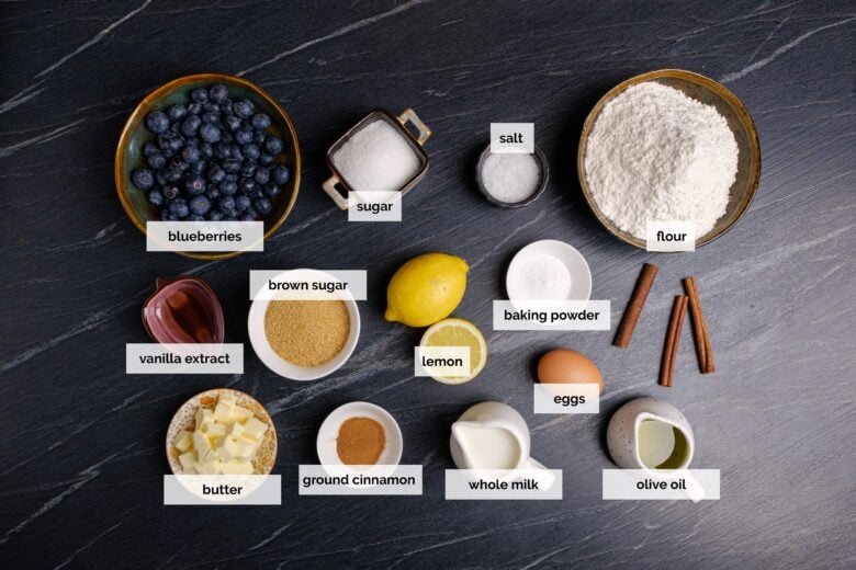 Overhead shot of ingredients to make bakery style blueberry muffins on a dark surface.