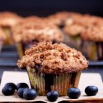 A close-up of a blueberry muffin with a crumbly topping on a slate surface, surrounded by scattered blueberries.