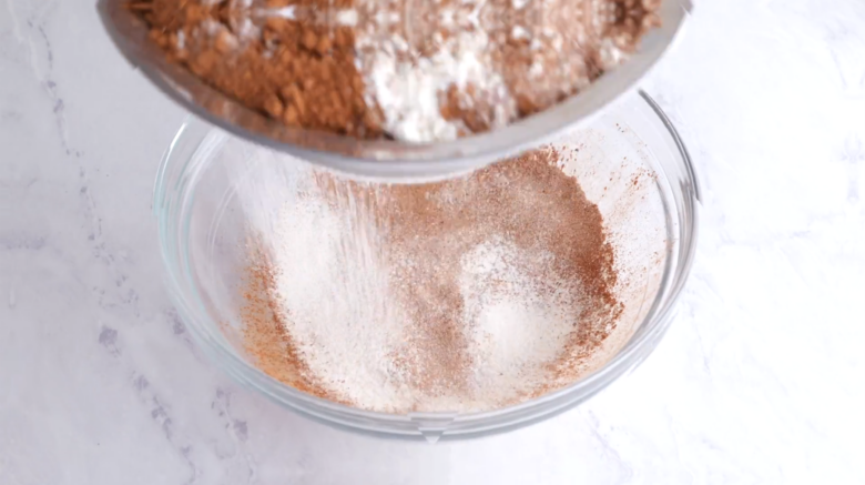 Flour, sugar, cocoa powder, and baking powder being sifted into a glass bowl to make Chocolate Strawberry Cake.