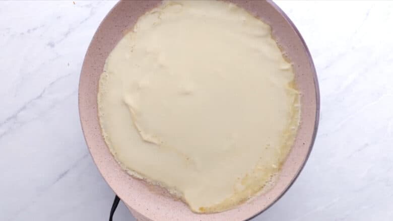 A crepe cooking in a pink non-stick frying pan on a stovetop.