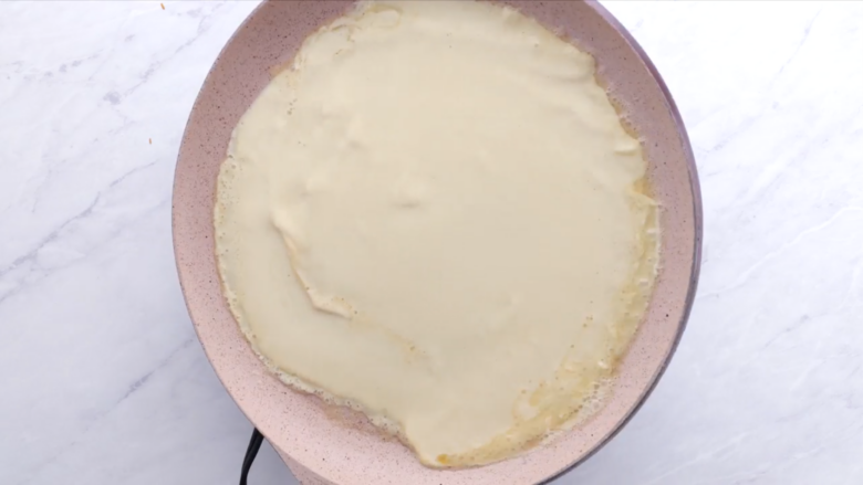 A crepe cooking in a pink non-stick frying pan on a stovetop.