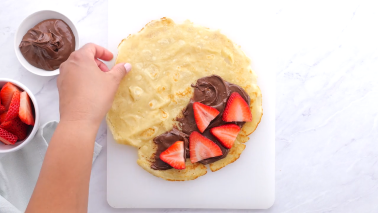 A crepe filled with Nutella and sliced strawberries on a cutting board.