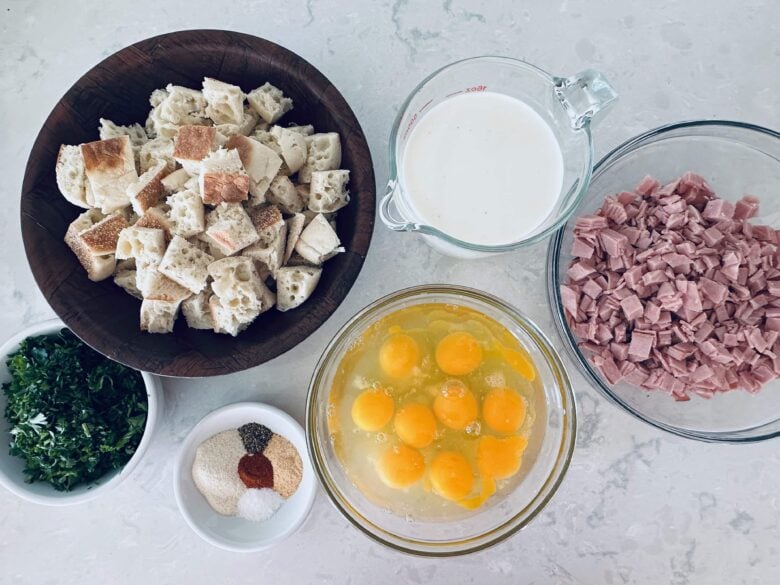 Overhead shot of ingredients to make eggs benedict casserole on a white marble countertop.