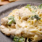 A plate of creamy chicken fettuccini alfredo pasta with broccoli and grated cheese on top, served on a ceramic plate.