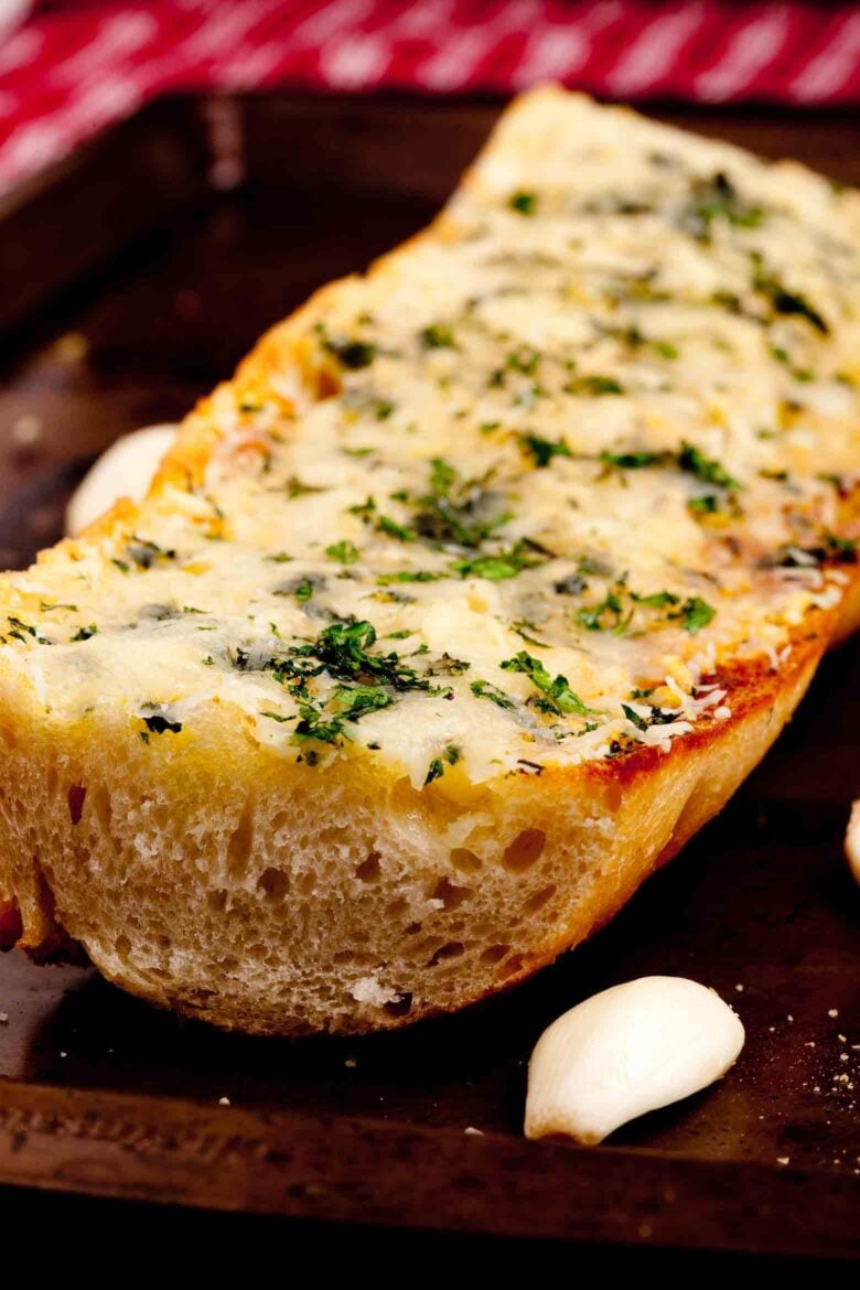 Close-up of a garlic bread topped with melted cheese and herbs, placed on a wooden board with whole garlic cloves beside it.