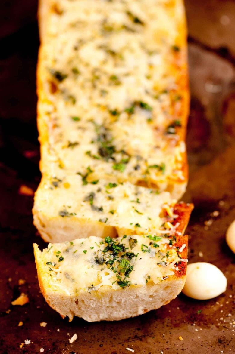 A close-up of a sliced loaf of garlic bread topped with melted cheese and herbs on a dark surface.