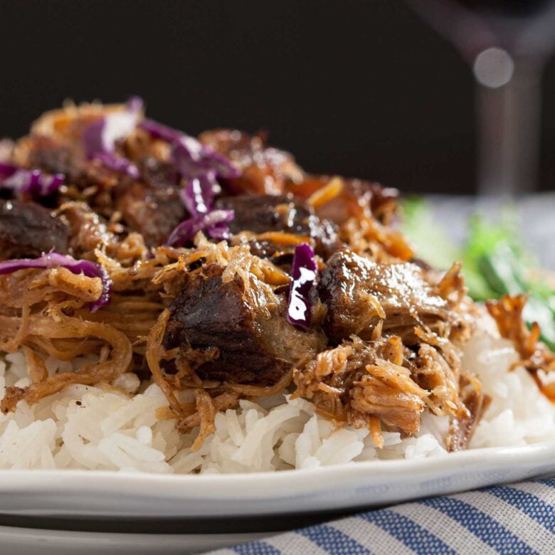 A close-up of a Kahlua Pork featuring pulled pork and purple cabbage served over white rice, with a glass of red wine in the background.