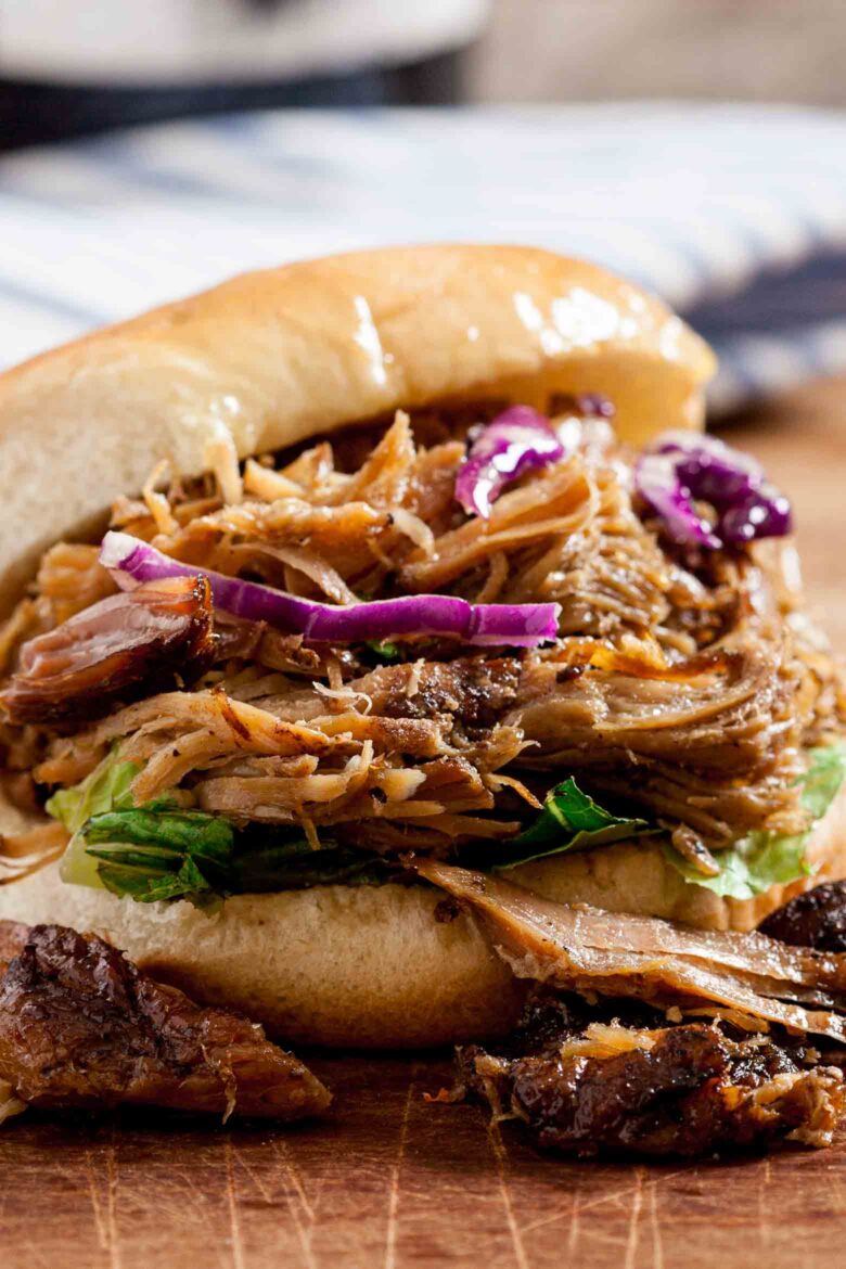 A pulled pork sandwich topped with coleslaw and red cabbage on a bun, served on a wooden table.