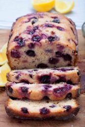 A lemon blueberry bread with three slices cut, showing visible berries. fresh lemon wedges are visible in the background.