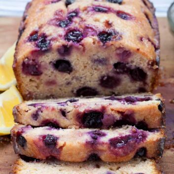 A lemon blueberry bread with three slices cut, showing visible berries. fresh lemon wedges are visible in the background.