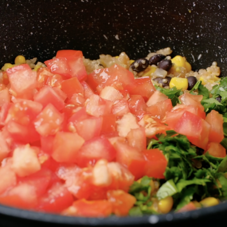Chopped tomatoes and cilantro being added to a pot with quinoa, corn, and black beans.