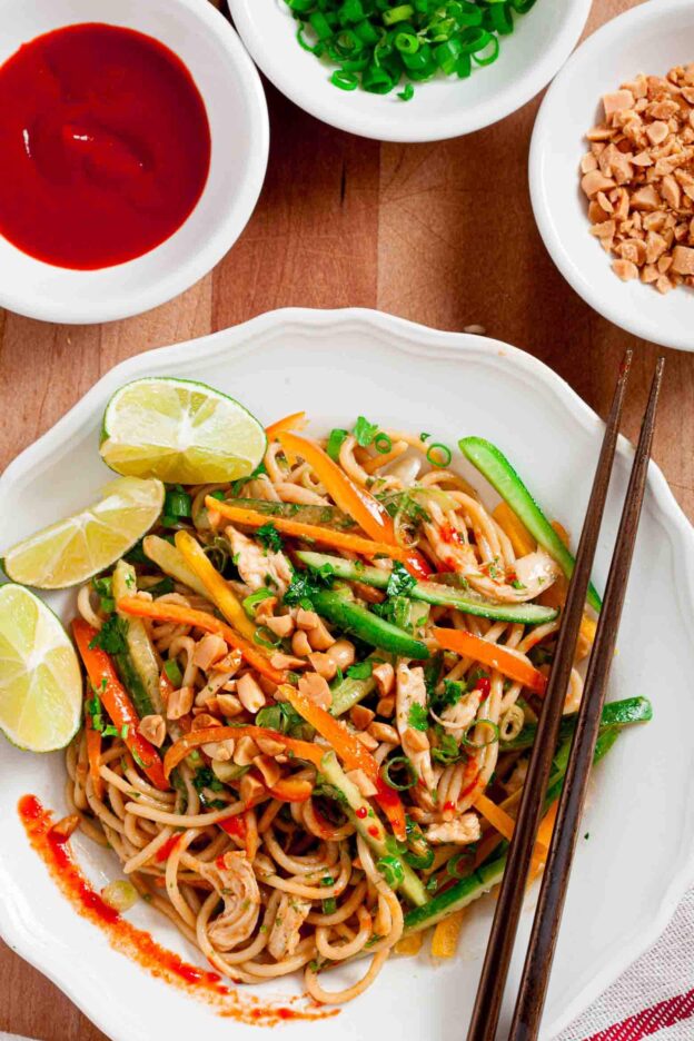 A bowl of Thai peanut noodles with vegetables, lime, and chili, accompanied by side dishes of sauce, chopped nuts, and spring onions.