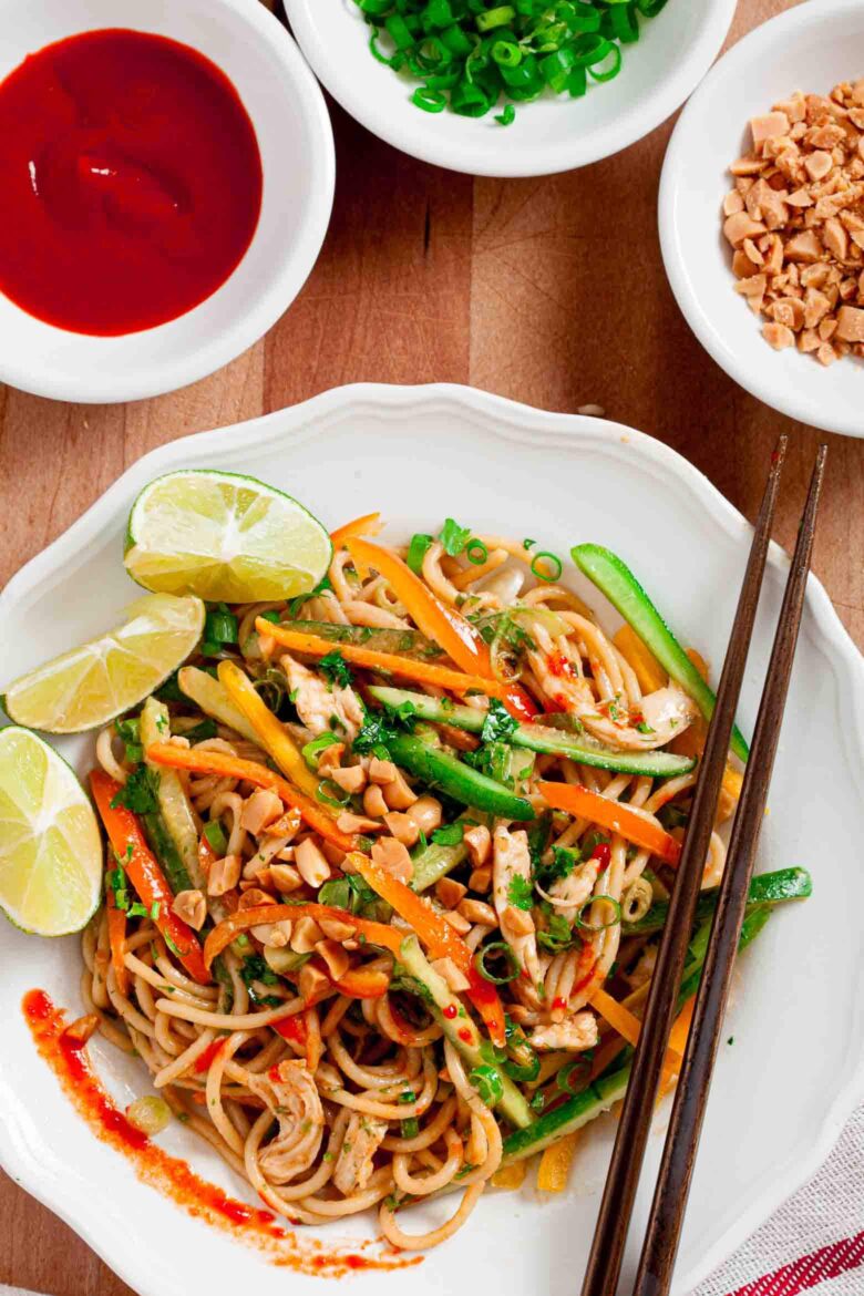 A bowl of Thai peanut noodles with vegetables, lime, and chili, accompanied by side dishes of sauce, chopped nuts, and spring onions.