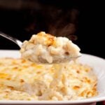 Closeup shot of a spoonful of steaming white cheddar mac and cheese being served from a baking dish with a golden crust.