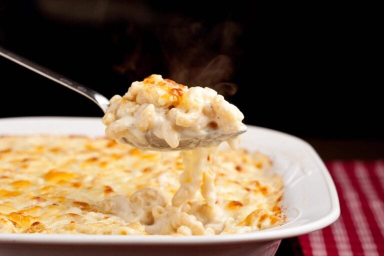 A steaming spoonful of baked macaroni and cheese is lifted from a dish, displaying a golden, bubbly top. The background is dark, highlighting the cheesy texture of the dish.