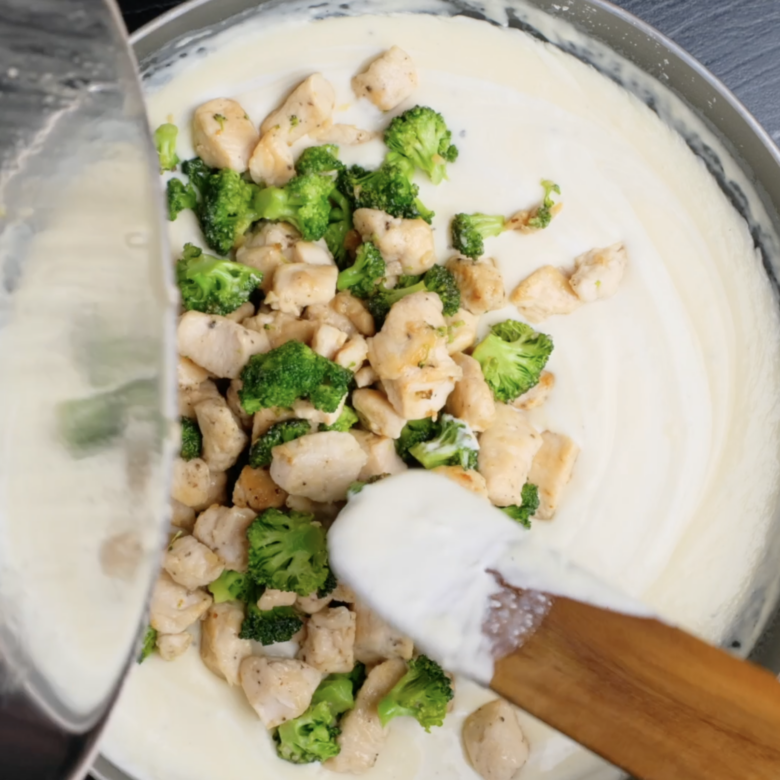 Cooked chicken and broccoli being added to Alfredo sauce in a pot.