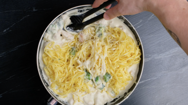 Cooked pasta being tossed into Alfredo sauce with chicken and broccoli.
