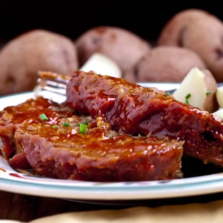 Glazed juicy meatloaf on a plate with potatoes in the background.