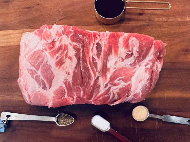 Raw pork shoulder on a wooden cutting board, surrounded by spices, a measuring spoon, and a cup of sauce.