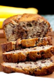 A sliced loaf moist chocolate chip banana bread on a wooden surface, with whole bananas visible in the background.