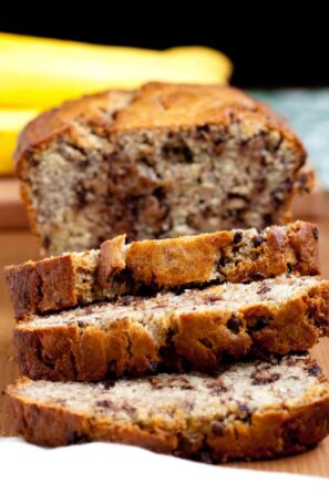 A sliced loaf moist chocolate chip banana bread on a wooden surface, with whole bananas visible in the background.