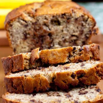 A sliced loaf moist chocolate chip banana bread on a wooden surface, with whole bananas visible in the background.