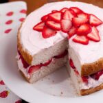 A sliced fresh strawberry cake on a white plate, topped with fresh strawberry slices.
