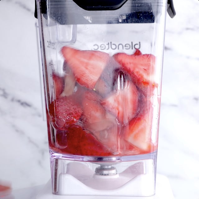 Close-up of a clear blender jar filled with chopped strawberries against a marble background.