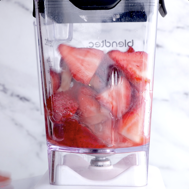 Close-up of a clear blender jar filled with chopped strawberries against a marble background.