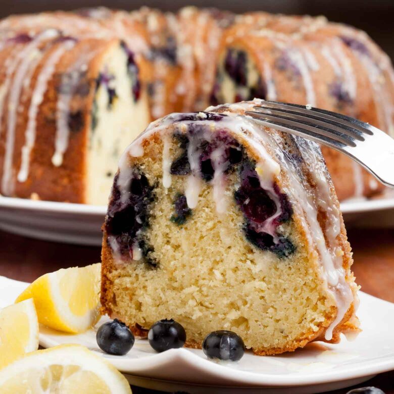 A slice of blueberry lemon bundt cake with icing on a plate, garnished with lemon slices and blueberries. 