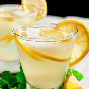 A close-up of a glass filled with a lemonade margarita, garnished with lemon slices, ice, and a sugared rim.