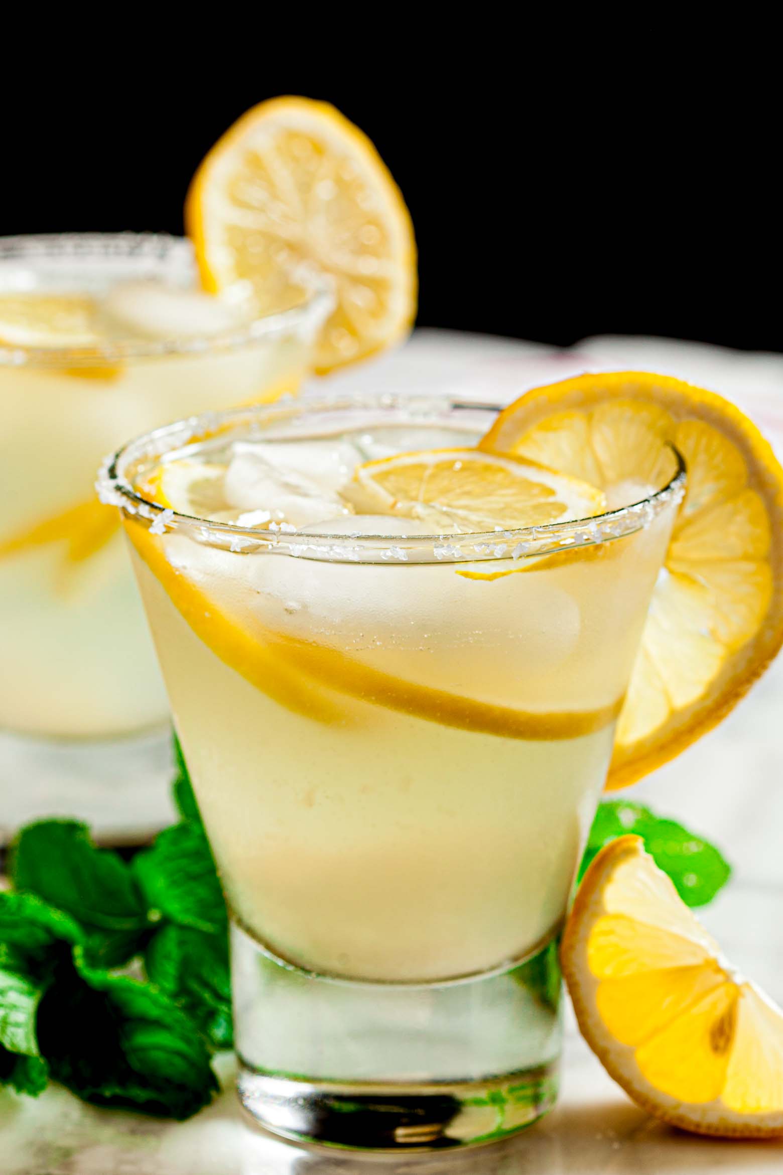 A close-up of a glass filled with a lemonade margarita, garnished with lemon slices, ice, and a sugared rim.