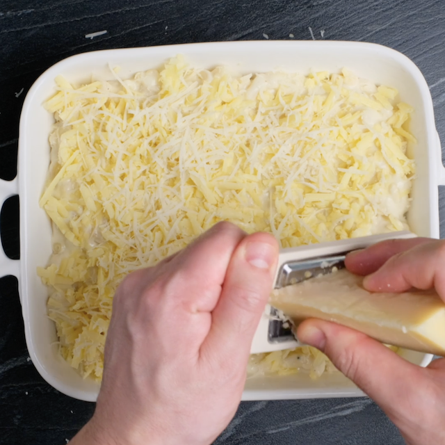 Grated cheese being added to assembled white cheddar mac and cheese in a baking pan.
