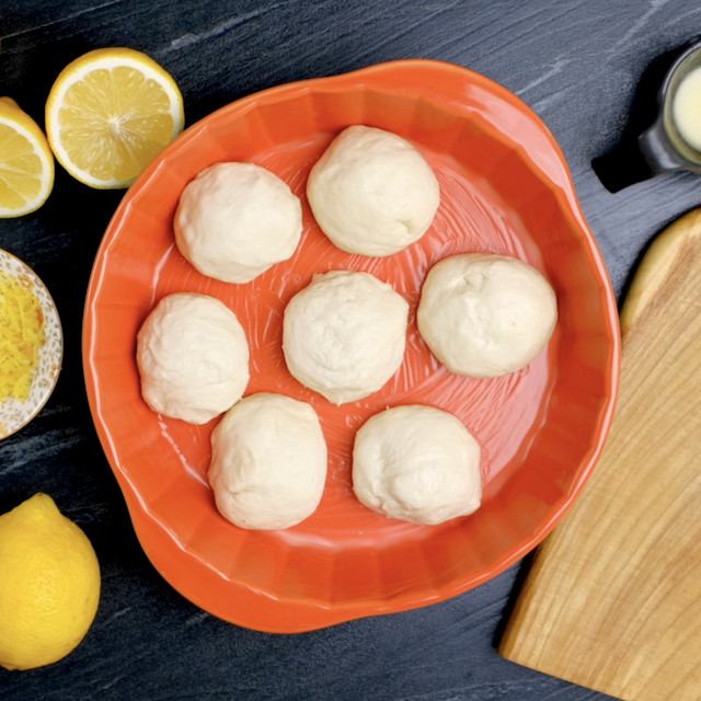 A red baking dish on a black surface contains seven dough balls.