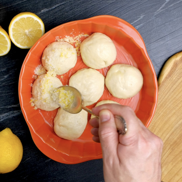 A hand sprinkles lemon zest and sugar mixture over seven round dough balls in a baking dish.