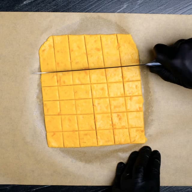 Rolled out cheese cracker dough being cut into squares using a sharp knife.