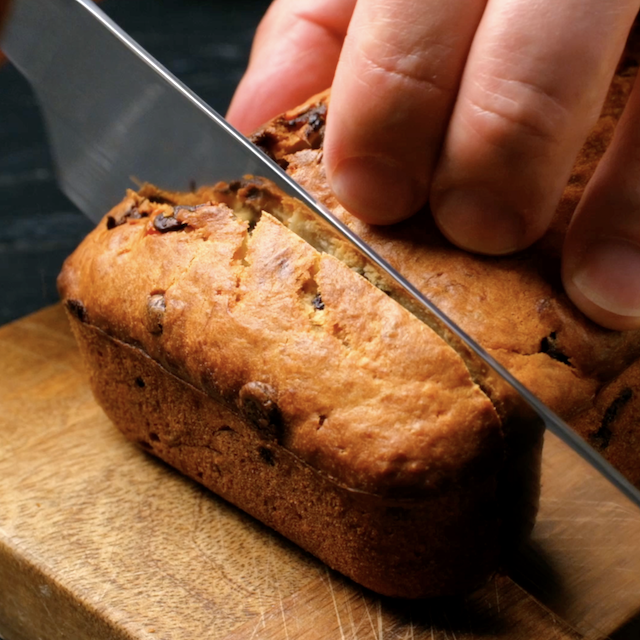A hand holds a knife cutting into a loaf of moist chocolate chip banana bread on a wooden cutting board.