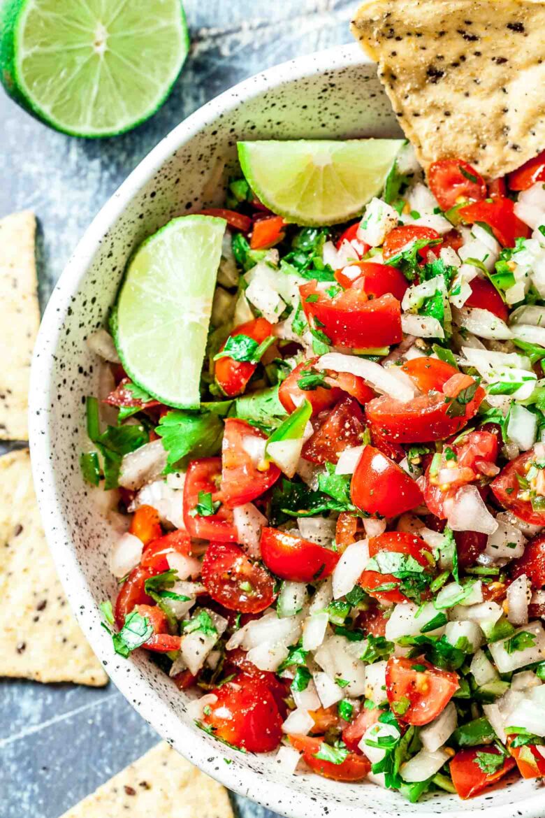 A bowl of fresh pico de gallo made with diced tomatoes, onions, cilantro, and lime wedges, accompanied by tortilla chips.
