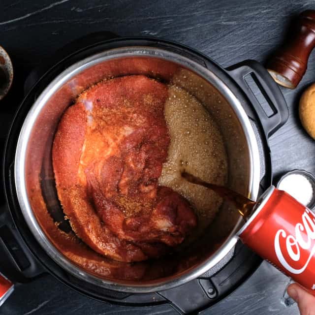 Coca cola being poured into a slow cooker with seasoned pork shoulder.