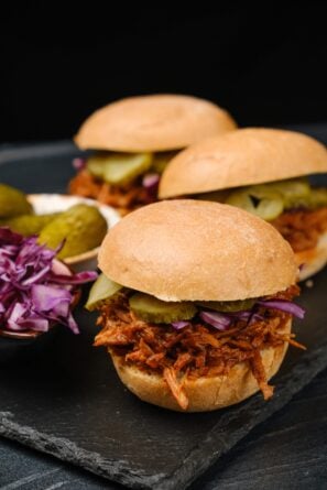 Three BBQ pulled pork sandwiches with pickles and shredded purple cabbage on a black slate serving board.