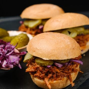 Three BBQ pulled pork sandwiches with pickles and shredded purple cabbage on a black slate serving board.